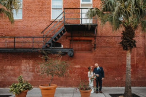 Ybor City Tampa Wedding Bride and Groom Outdoor Brick Wall Portrait | Long Sleeve Lace Bridal Gown Wedding Dress | Classic Navy Blue Groom Suit | Organic Natural Loose Wedding Bridal Bouquet with Coral Pink Peonies and White Roses with Succulents and Eucalyptus Greenery by Tampa Florida Monarch Events and Designs
