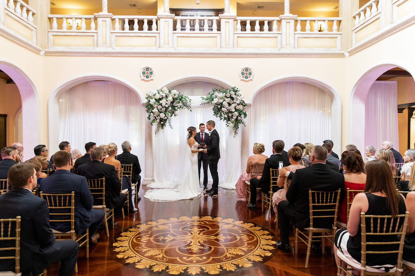 Bride and Groom Exchanging Vows at Tampa Wedding Venue Avila Golf & Country Club Indoor Ceremony with Gold Chiavari Chairs, Pipe and Drape Backdrop with Blush Pink and Ivory Rose Floral Arrangements with Eucalyptus Greenery