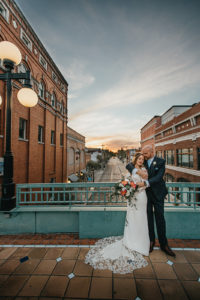Ybor City Tampa Wedding Bride and Groom Outdoor Historic Building Portrait | Long Sleeve Lace Bridal Gown Wedding Dress | Classic Navy Blue Groom Suit | Organic Natural Loose Wedding Bridal Bouquet with Coral Pink Peonies and White Roses with Succulents and Eucalyptus Greenery by Tampa Florida Monarch Events and Designs