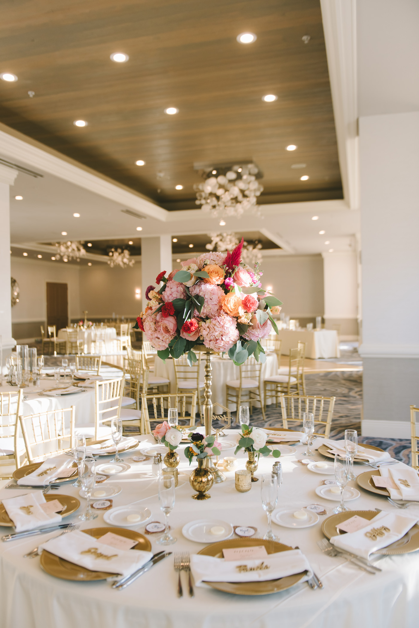 Clearwater Wedding Venue The Hyatt Regency Clearwater Beach Reception Ballroom with White Table Linens and Gold Chiavari Chairs and Gold Charger Plates | Vibrant Bright Colorful Tall Centerpieces with Pink Hydrangea and Orange Roses atop Gold Candlesticks | Gabro Event Services Wedding Rentals
