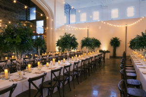 Wes Anderson Inspired Wedding Reception Decor, Long Feasting Tables with Wooden Cross back Chairs, Tall Greenery Plants Centerpieces and String Lights, Hurricane Glass Candles | Wedding Photographer Carrie Wildes Photography | Tampa Bay Wedding Venue The Rialto Theatre | Wedding Planner UNIQUE Weddings and Events | Wedding Florist Monarch Events and Design | Wedding Caterer Olympia Catering