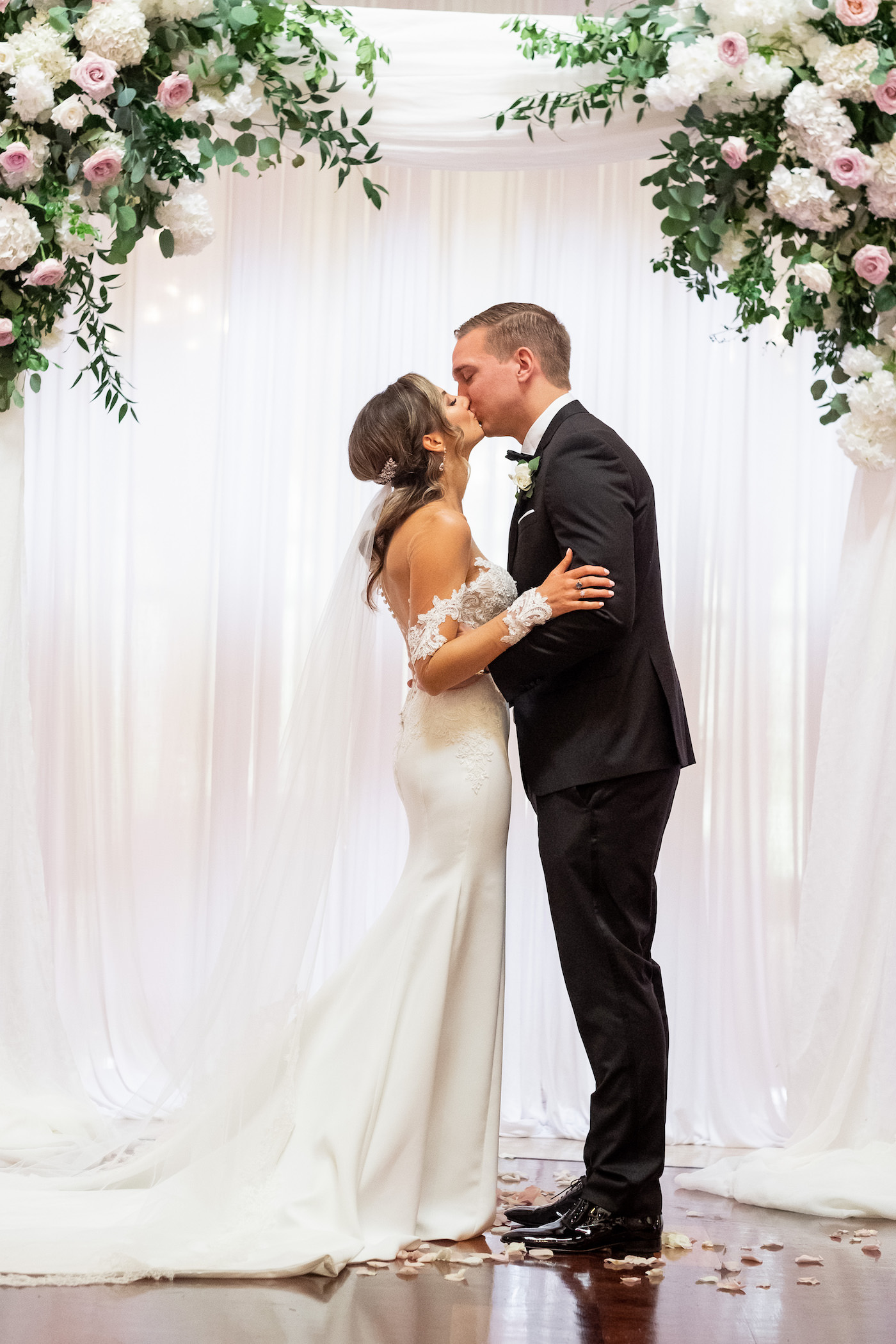 Bride and Groom First Kiss Bride and Groom Exchanging Vows during Indoor Ceremony with Pipe and Drape Backdrop with Blush Pink and Ivory Rose Floral Arrangements with Eucalyptus Greenery