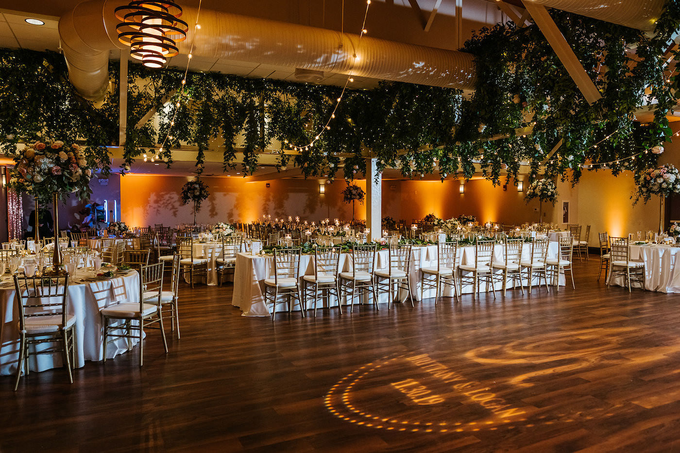 St. Pete Garden Wedding Indoor Reception with Amber Gold Uplighting and Wedding Ceiling Floral Installment with Greenery and Blush Pink and White Roses and Canopy String Lights | Monogram Dance Floor Gobo | Reception Table with White Linens and Gold Chiavari Chairs | Tampa Wedding Rentals A Chair Affair