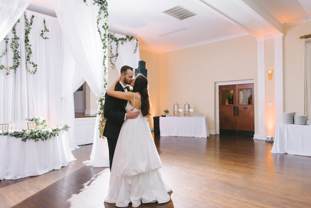 Romantic Classic Bride and Groom Kissing During First Dance Wedding Reception Ballroom Portrait | White Linen Draping with Lush Greenery Garland and Sweetheart Table | Tampa Bay Wedding Photographer Kera Photography | Wedding Planner Breezin' Weddings | Historic Wedding Venue The Orlo | Wedding DJ Grant Hemond and Associates