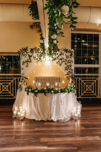 Sweetheart Table with Draped Arch Backdrop with Cascading Greenery and Candles and Loveseat for Bride and Groom | Tampa Wedding Rentals A Chair Affair