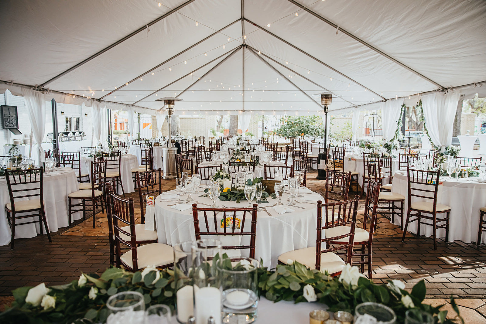 Outdoor Garden Wedding Reception Tent with Canopy String Lights | Round White Tables with Mahogany Wood Chiavari Chairs and Greenery Wreath Centerpieces with Lemons and Hurricane Pillar Candles by Tampa Wedding Florist Monarch Events and Designs