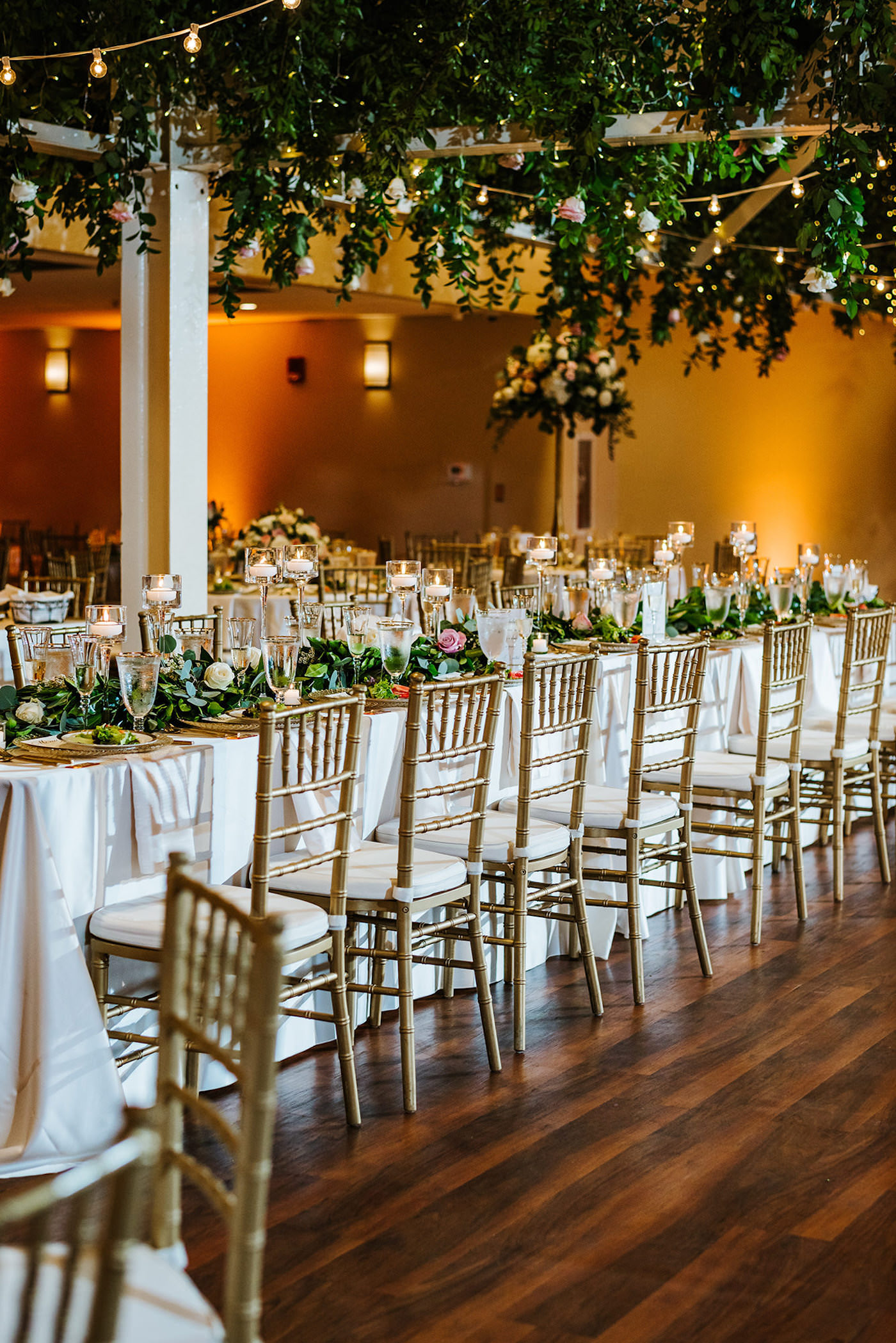 St. Pete Garden Wedding Indoor Reception with Amber Gold Uplighting and Wedding Ceiling Floral Installment with Greenery and Blush Pink and White Roses and Canopy String Lights | Reception Feasting Banquet Table with White Linens and Gold Chiavari Chairs | Monarch Events and Design | A Chair Affair