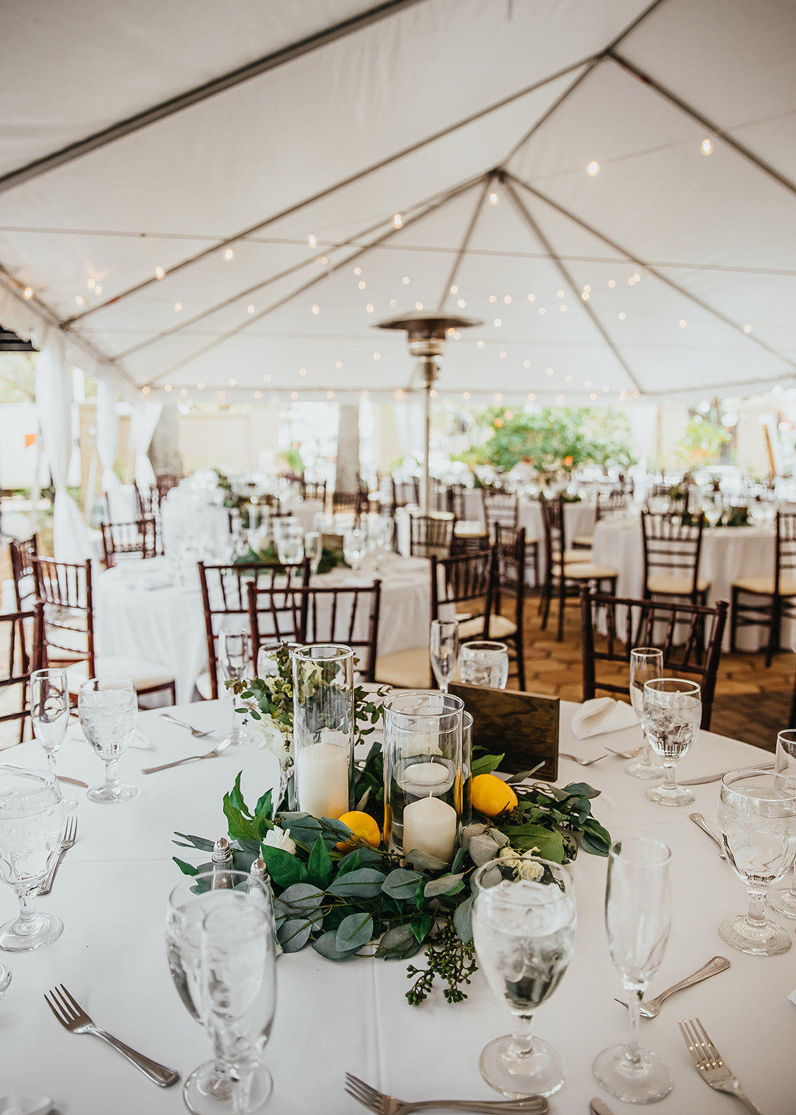 Outdoor Garden Wedding Reception Tent with Canopy String Lights | Round White Tables with Mahogany Wood Chiavari Chairs and Greenery Wreath Centerpieces with Lemons and Hurricane Pillar Candles by Tampa Wedding Florist Monarch Events and Designs