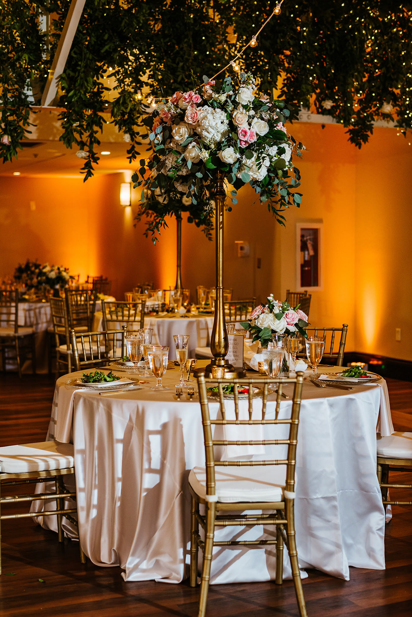 Tall Wedding Centerpiece with White Hydrangea and Blush Pink Roses and Eucalyptus Greenery on top of Gold Candlestick | St. Pete Garden Wedding Indoor Reception with Amber Gold Uplighting and Wedding Ceiling Floral Installment with Greenery and Blush Pink and White Roses and Canopy String Lights | Reception Table with White Linens and Gold Chiavari Chairs | Monarch Events and Design | A Chair Affair