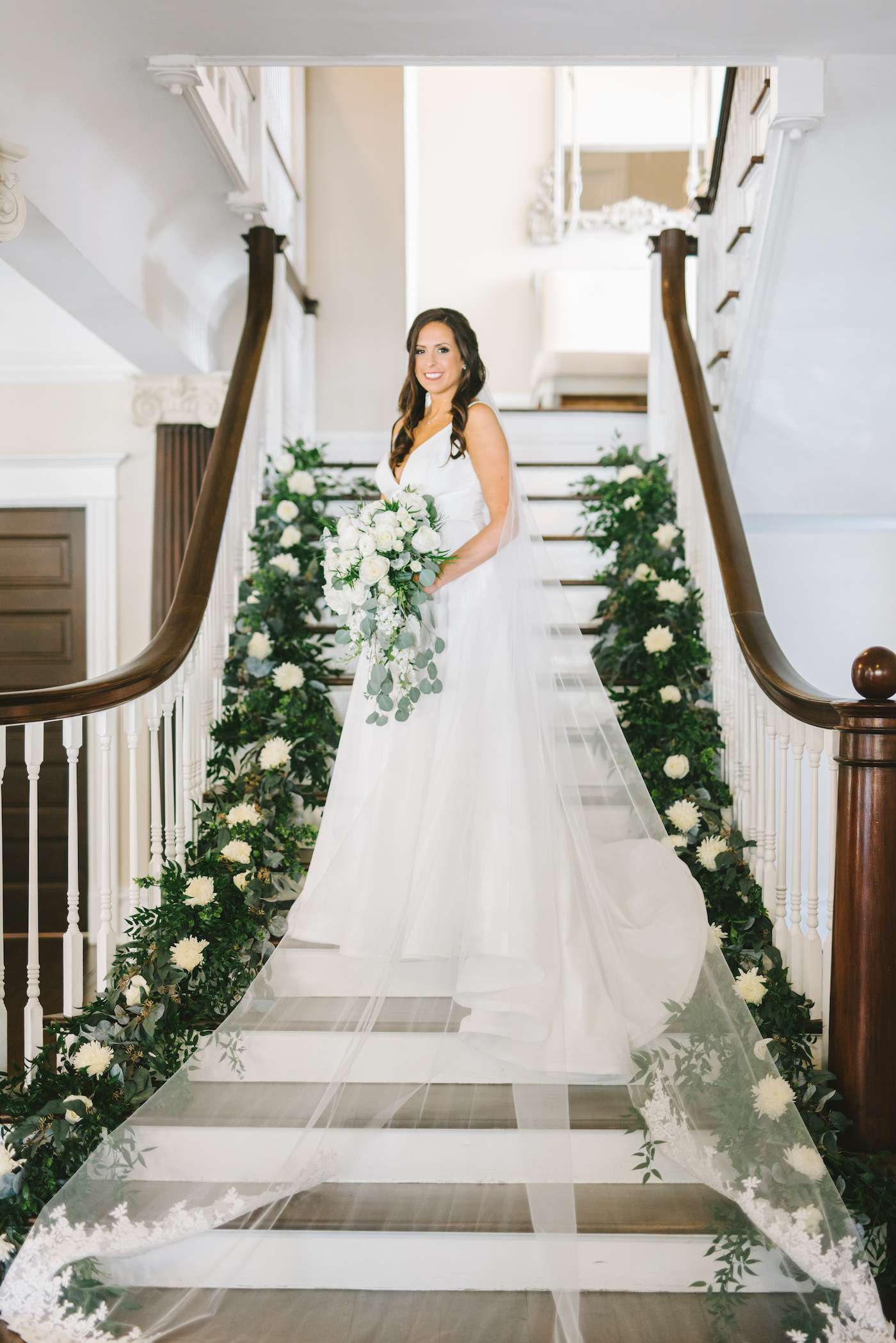 Romantic Classic Bride in A-Line White Wedding Dress and Full Length Romantic Lace Trim Veil on Staircase Holding White Floral and Greenery Bridal Bouquet, Stairs Decorated with Lush Flowers | Tampa Bay Wedding Photographer Kera Photography | Tampa Bay Wedding Planner Breezin' Weddings | Historic Wedding Venue The Orlo