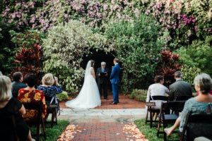 Bride and Groom Exchanging Vows in Outdoor Whimsical St. Petersburg Wedding Venue Sunken Gardens with Greenery and Purple Flower Backdrop | Tampa Wedding Planner Kelly Kennedy Weddings and Events