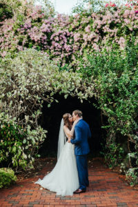 Intimate Kissing Portrait of Bride and Groom in Front of Greenery and Purple Lilac Natural Floral Backdrop | St. Pete Wedding Venue Sunken Gardens