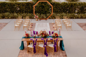 Boho Glam Wedding Ceremony Backdrop with Wood Geometric Arch and Gold Chiavari Chairs and Rug Runner | Colorful Orange Pink and Red Floral Centerpiece with Pampas Grass | Tampa Wedding Florist Monarch Events and Designs | Wood Farm Table with Teal Turquoise Runner and Colorful Taper Candles and Purple Chair Sashes