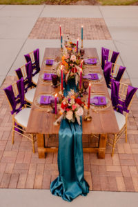 Boho Glam Wedding Reception Table with Orange Pink and Red Floral Arrangement Centerpiece with Pampas Grass | Tampa Wedding Florist Monarch Events and Designs | Wood Farm Table with Teal Turquoise Runner and Colorful Taper Candles and Purple Sashes on Gold Chiavari Chairs