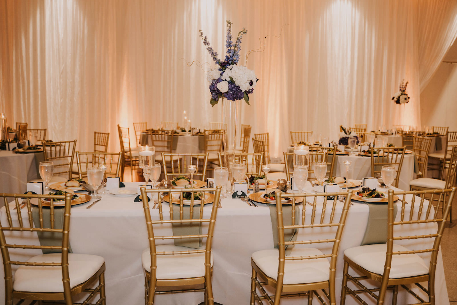 Tampa Wedding Venue the Tampa Garden Club Indoor Reception | Pipe and Drape with Uplights | Reception Tables with White Linens and Gold Chiavari Chairs and Gold Charger Plates with Tall Clear Vase Centerpieces of Purple and White Hydrangea Roses and Stock Flowers