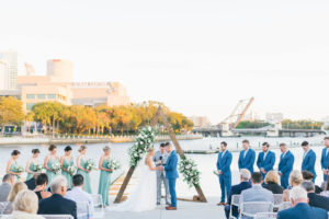 Boho Chic Waterfront Wedding Ceremony Portrait of Bride and Groom Exchanging Wedding Vows, Wooden Triangular Arch with Lush Greenery and Ivory, White Roses, Bridesmaids in Green Dresses | Wedding Venue Tampa River Center | Wedding Florist Bruce Wayne Florals | Wedding Planner Parties A'la Carte