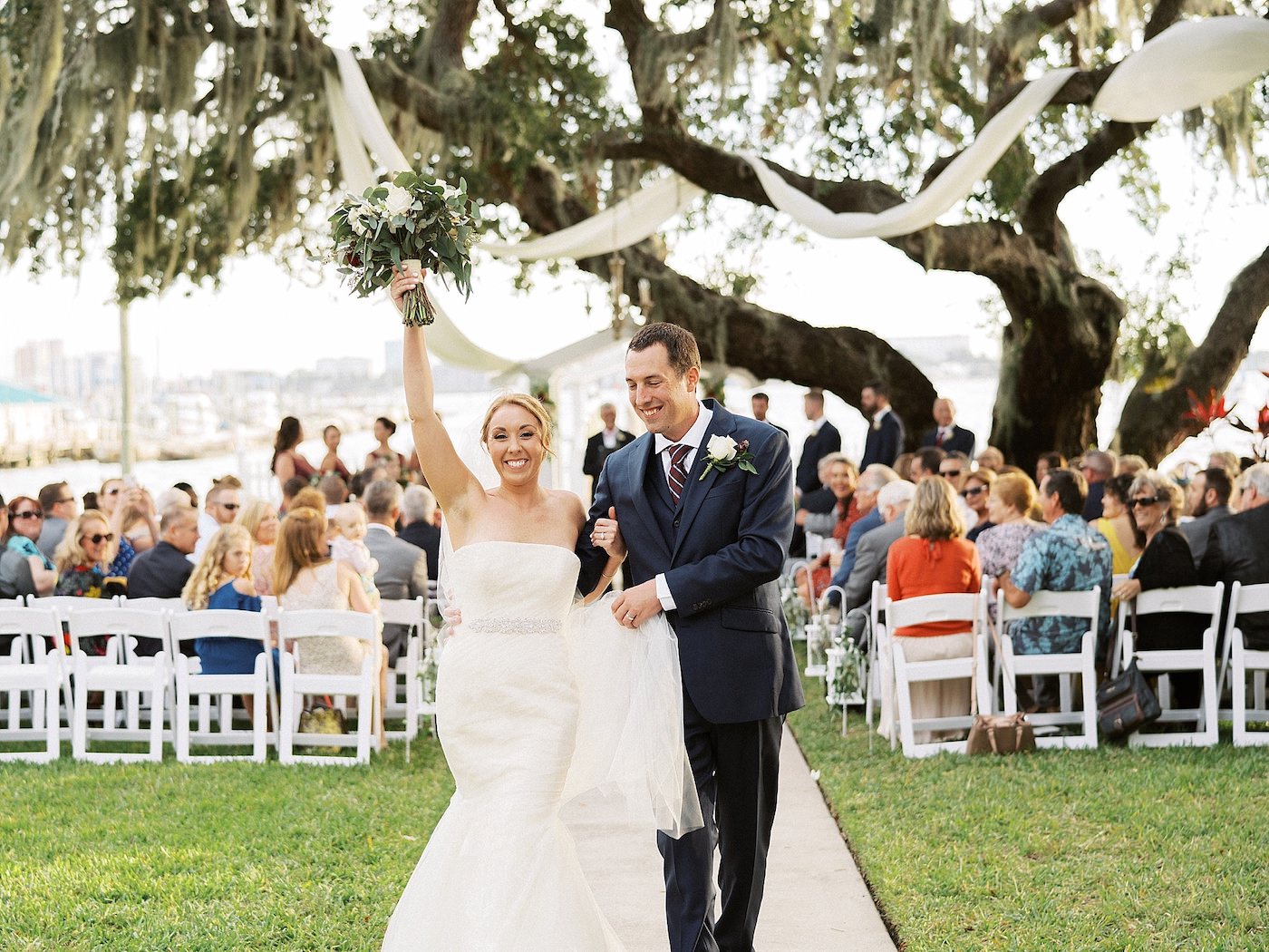 Bride and Groom Walking Down the Aisle | Florida Fall Autumn Outdoor Waterfront Wedding Ceremony | Strapless Ivory Tulle Rouched Mermaid Vera Wang Bridal Gown with Rhinestone Beaded Sash Waistband | Groom in Classic Navy Blue Suit
