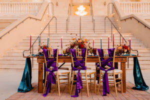 Boho Glam Wedding Reception Table with Orange Pink and Red Floral Arrangement Centerpiece with Pampas Grass | Tampa Wedding Florist Monarch Events and Designs | Wood Farm Table with Teal Turquoise Runner and Colorful Taper Candles and Purple Sashes on Gold Chiavari Chairs | Tampa Wedding Venue The Regent Grand Staircase and Columns