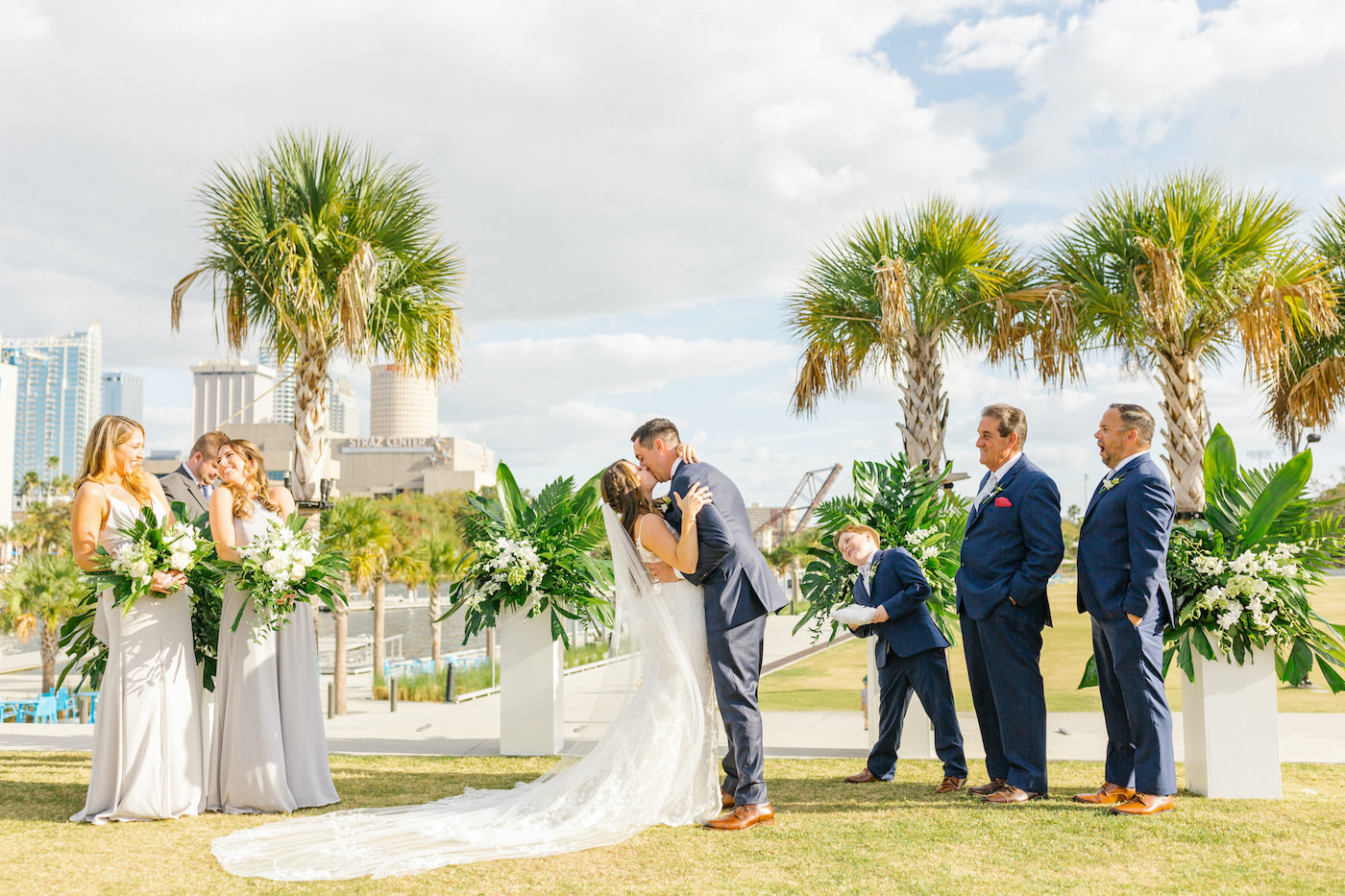 Bride and Groom First Kiss during Tropical Florida Wedding Outdoor Downtown Tampa Ceremony | White and Green Ceremony Floral Arrangements with Palm Leaves and Ferns and White Orchids atop White Columns