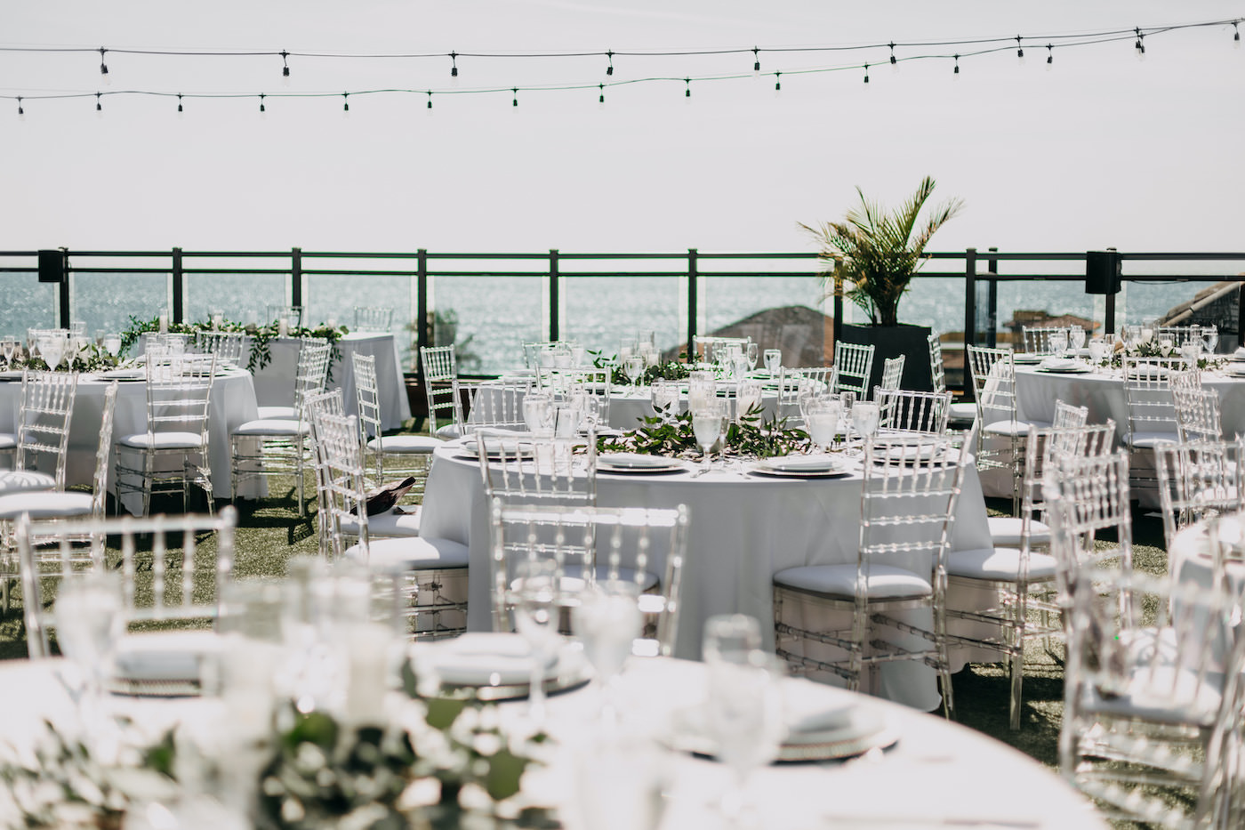 Florida Waterfront Rooftop Reception | Round Tables with White Linens, Clear Acrylic Ghost Chairs, Low Floral Centerpieces with Greenery | Gulf Coast Boutique Hotel and Wedding Venue The Hotel Zamora | St. Pete Beach Wedding Planner Blue Skies Weddings and Events | Tampa Bay Rental Company Gabro Event Services | Wedding DJ Grant Hemond and Associates