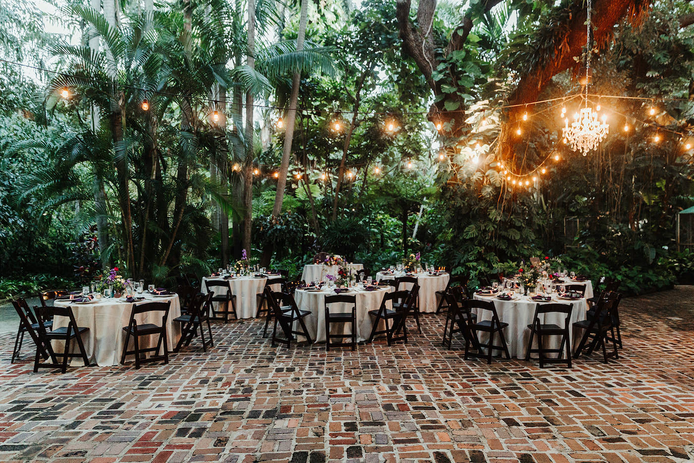 Whimsical Boho Outdoor Courtyard Garden Wedding Reception Decor, Round Tables with Wooden Folding Chairs, Cream Linens, Colorful Floral Centerpieces, Hanging Crystal Chandelier and String Lights | Tampa Bay Wedding Planner Kelly Kennedy Weddings and Events | St. Pete Wedding Venue Sunken Gardens