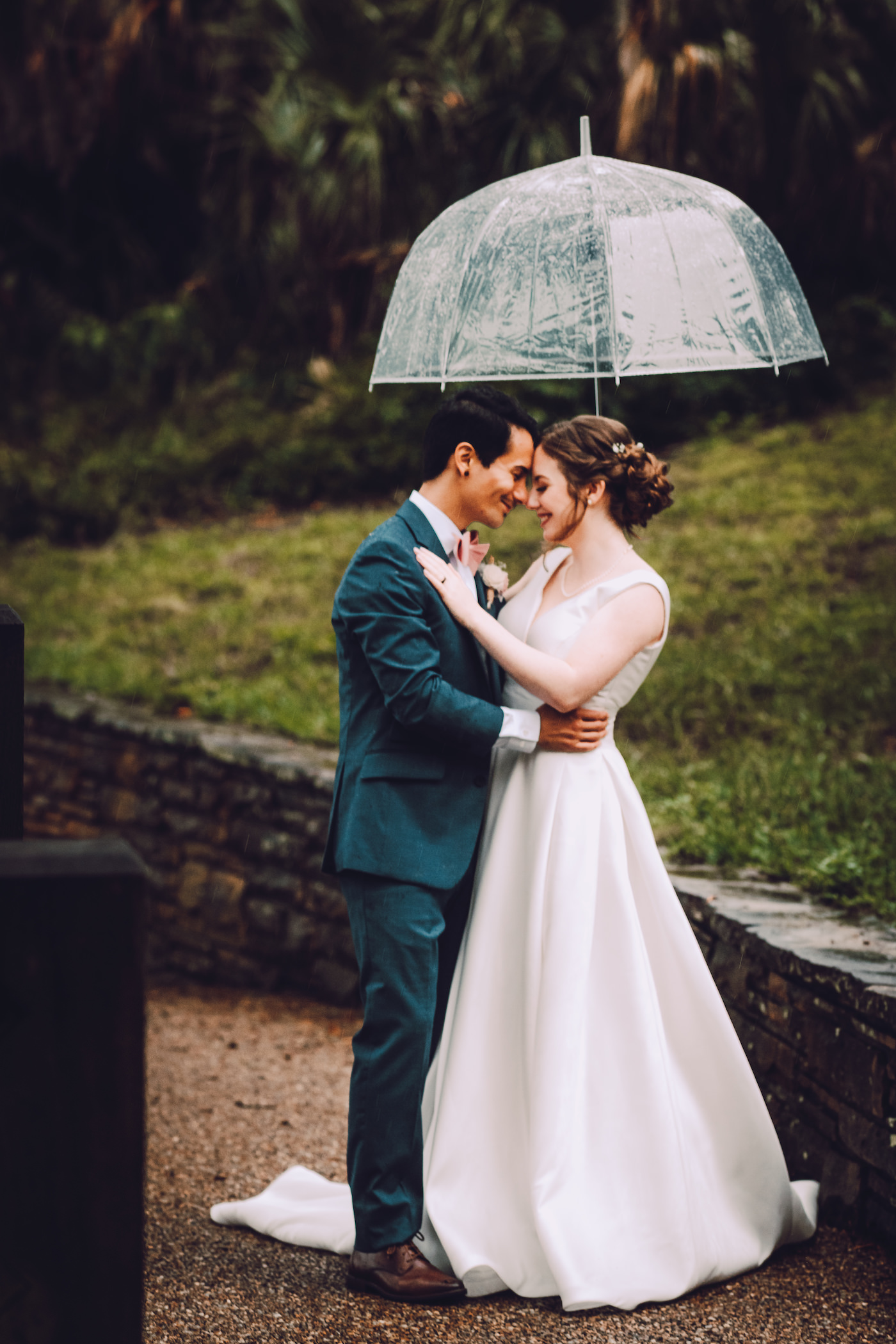 Bride and Groom Rainy Wedding Day Portrait with Clear Umbrellas | Stella York Satin V Neck Simple Classic Low Back Bridal Gown A Line Ballgown | Navy Blue Suit Groom Suit with Grey Vest and Blush Pink Bow Tie