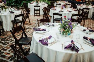 Whimsical Boho Outdoor Courtyard Wedding Reception Decor, Round Tables with Ivory Linens, Gold Beaded Rim Chargers, Purple Napkins, Colorful Wildflower Centerpiece