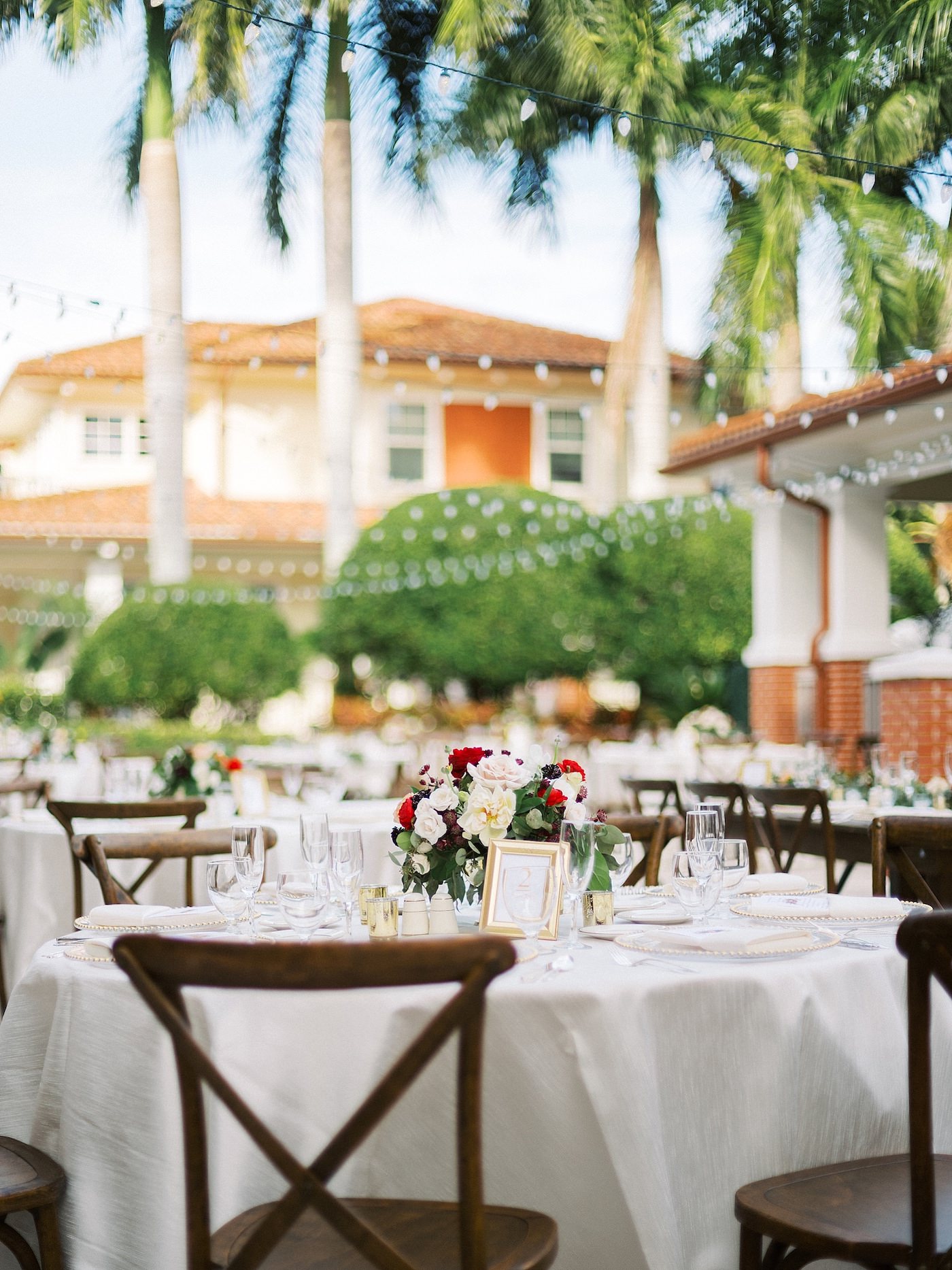 Florida Outdoor Wedding Reception with Canopy String Lights and Wood Cross Back French Country Chairs | Ivory Table Linens and Gold Frame Table Numbers with Low Floral Centerpiece of Red and White Roses and Greenery