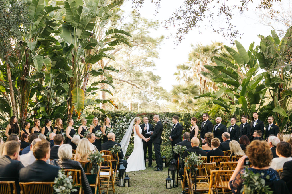 Bride and Groom Exchanging Vows during Boho Chic Outdoor Wedding Ceremony at Tampa Garden Club Venue | Natural Wood Bamboo Chairs with Black Lanterns and Eucalyptus Greenery Aisle Markers | Round Wood Moon Arch Ceremony Backdrop with Suspended Geometric Containers and Eucalyptus Greenery Garland Floral Arrangements | Black and White Wedding with Bridesmaids in Black Dresses and Groomsmen in Black Tux Suit