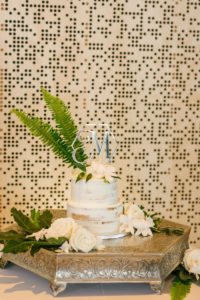 Simple Tropical Wedding Cake | Two Tier Half Naked Buttercream Wedding Cake with Fresh Flowers of White Roses and Orchids and Fern Greenery topped with Silver Monogram Cake Topper