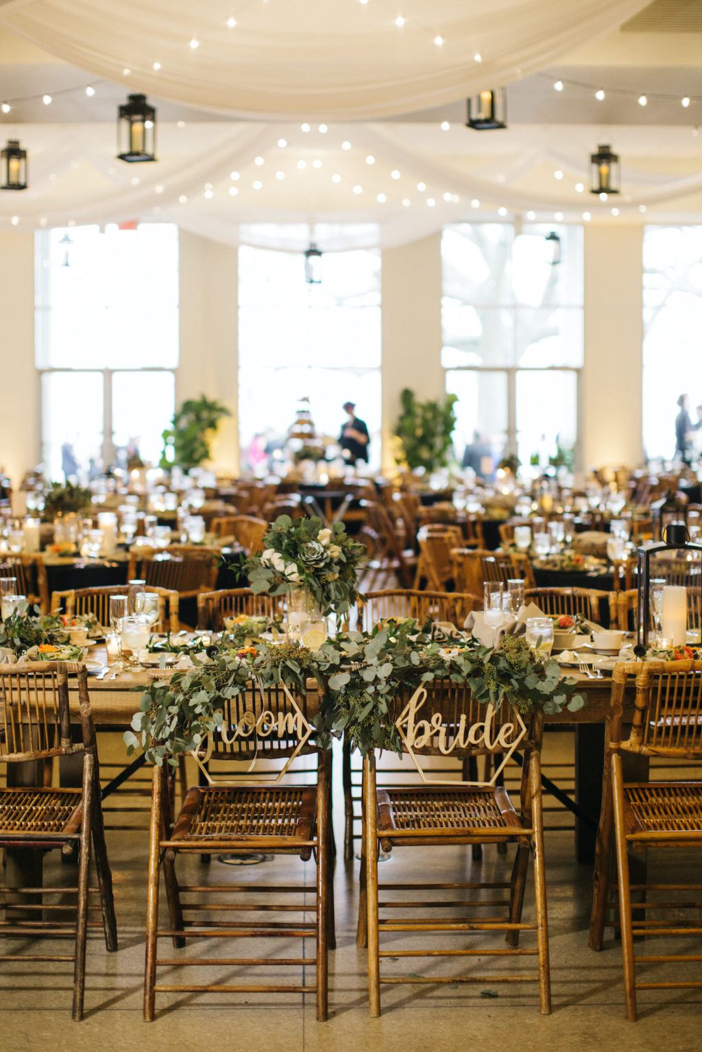 Bride and Groom Gold Geometric Chair Signs with Eucalyptus Greenery Garland | Boho Chic Wedding Reception at Tampa Wedding Venue the Tampa Garden Club | Wedding Ceiling Treatment Installment with Draped Fabric Swags and Canopy Bistro Lights and Suspended Black Lanterns with LED Candles | Tampa Wedding Rentals Gabro Event Services | Black and White Wedding with Natural Wood Bamboo Chairs and Wood Farm Feasting Tables and Black Table Linens with Black Lantern and Candle Centerpieces | Winsor Event Studio