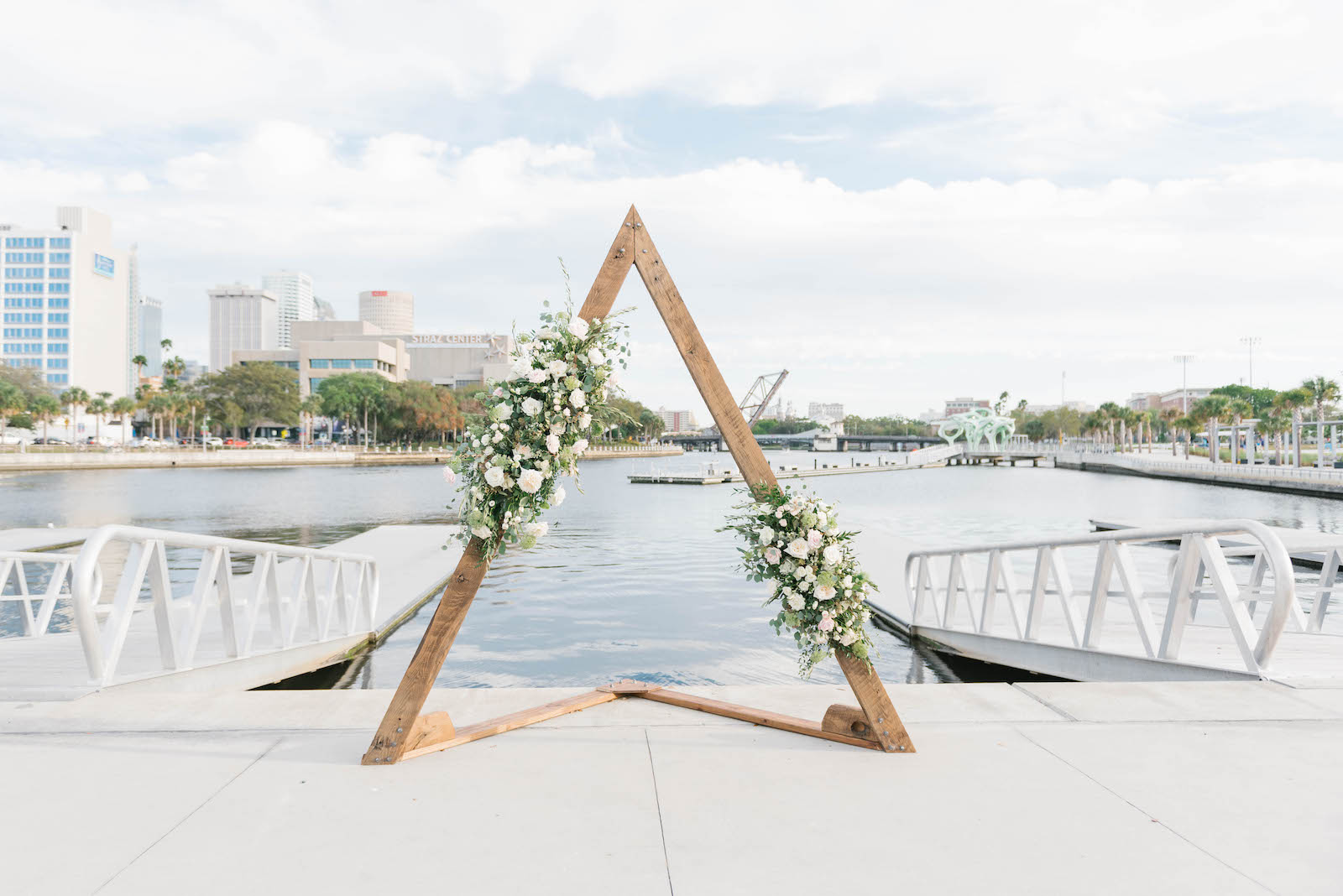 Boho Chic Waterfront Wedding Ceremony Decor, Triangular Wooden Arch with Lush Greenery and White, Ivory Roses | Wedding Venue The Tampa River Center | Wedding Venue Parties A'la Carte | Wedding Florist Bruce Wayne Florals
