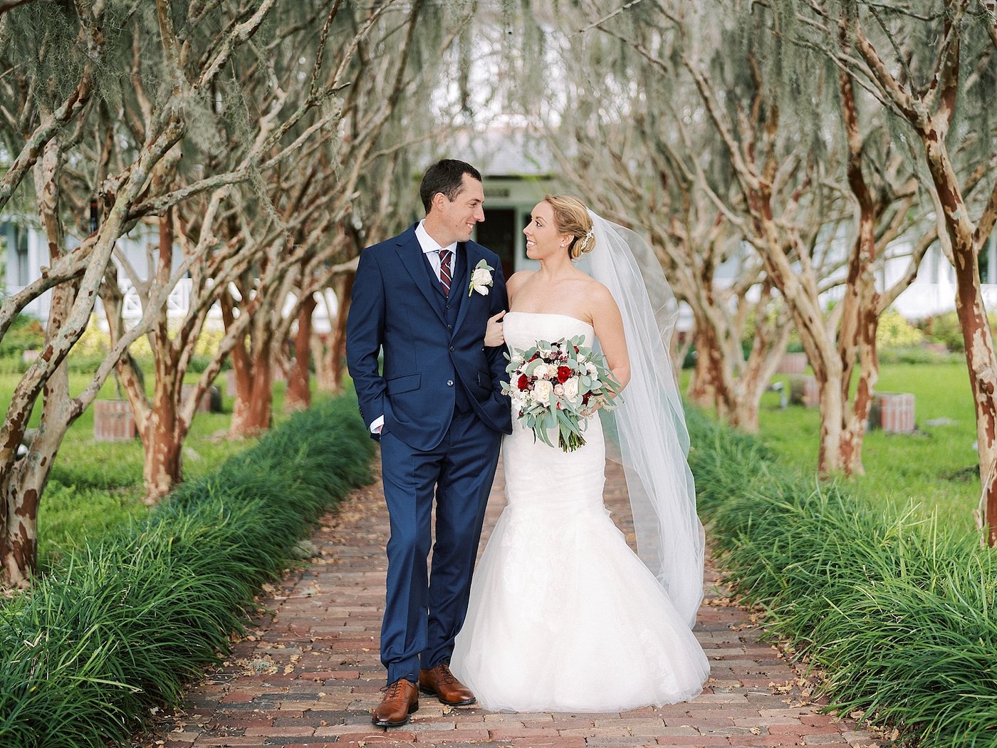 Bride and Groom Outdoor Portrait in Tree Lined Path | Strapless Ivory Tulle Rouched Mermaid Vera Wang Bridal Gown with Rhinestone Beaded Sash Waistband | Burgundy Wine Bordeaux Red and White Bridal Bouquet with Eucalyptus Greenery by Tampa Wedding Florist Brides N Blooms | Groom in Classic Navy Blue Suit