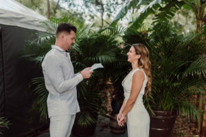 Bride and Groom Exchanging Vows during COVID Wedding Elopement Backyard Ceremony | Ivory White Bridal Jumpsuit | Casual Groom White Shirt and Pants