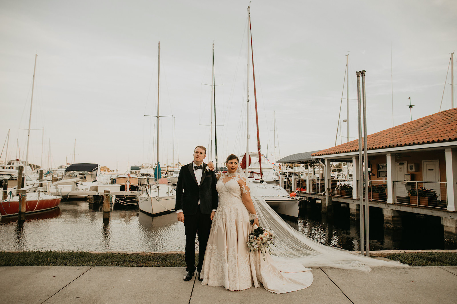 Downtown St. Petersburg Florida Marina Outdoor Bride and Groom Portrait | Champagne and Ivory Lace Allure Bridal Ball Gown with Sweetheart Neckline and Illusion Lace Bodice and Sleeves with Cathedral Veil | Groom Wearing Classic Black Tuxedo Suit with Bow Tie