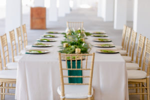 Clearwater Beach Wedding Venue Hilton Clearwater Beach | Modern Tropical Beach Outdoor Wedding Reception Terrace Feasting Table with Champagne Table Linens and Emerald Green Napkins under Gold Charger Plates | Gold Chiavari Chairs and Tropical Palm Frond Leaf Floral Arrangement Garland