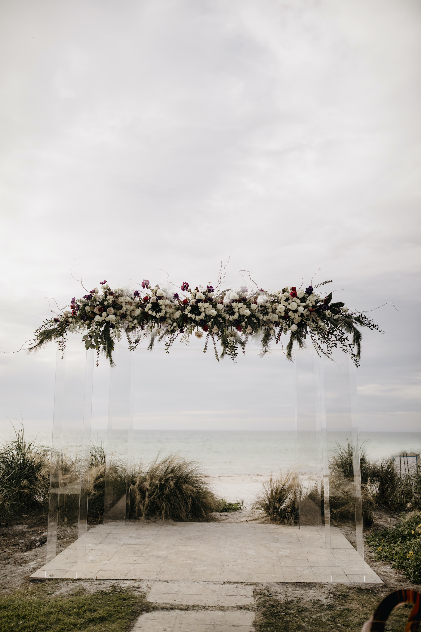 Sarasota Outdoor Beach Wedding Ceremony on Siesta Key | Acrylic Four Post Arbor Backdrop with White and Deep Red and Blush Pink Floral Arrangement Garland with Eucalyptus Greenery