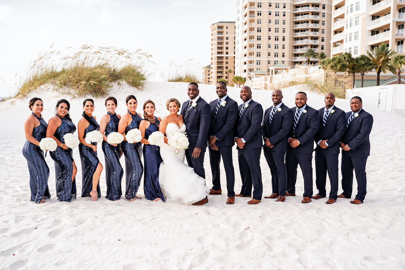 Romantic Tropical Florida Wedding Party, Bride and Groom Beachfront Portraits, Bridesmaids Wearing Matching Sophisticated Navy Sequined Halter Dresses, Bride Fit and Flare Strapless Wedding Dress Holding Ivory and White Round Floral Bouquet | Florida Gulf of Mexico Hotel and Wedding Venue Hilton Clearwater Beach