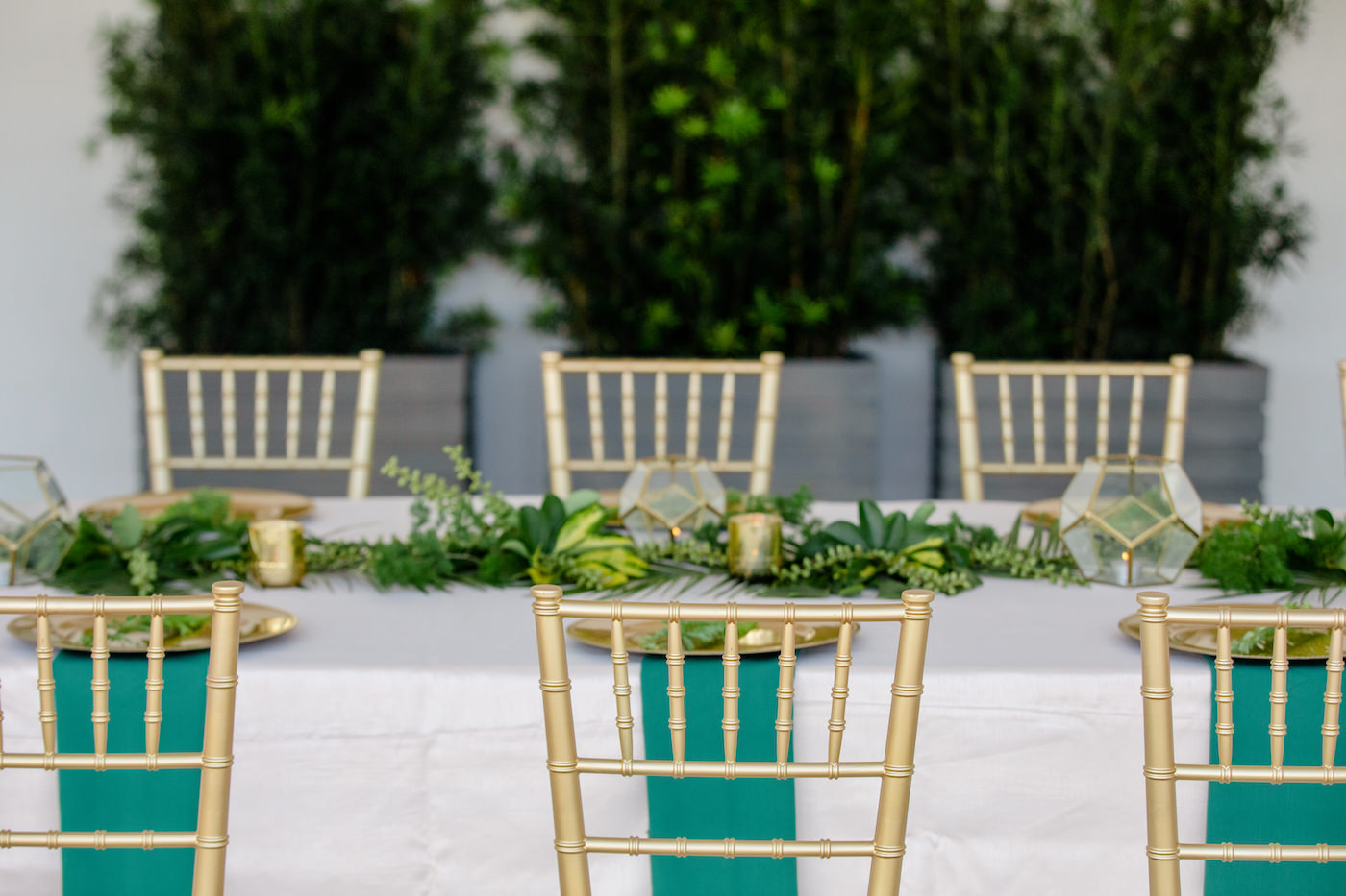 Clearwater Beach Wedding Venue Hilton Clearwater Beach | Modern Tropical Beach Outdoor Wedding Reception Terrace Feasting Table with Champagne Table Linens and Emerald Green Napkins under Gold Charger Plates topped with Ferns | Gold Chiavari Chairs and Tropical Palm Frond Leaf Floral Arrangement Garland and Gold Geometric Candles