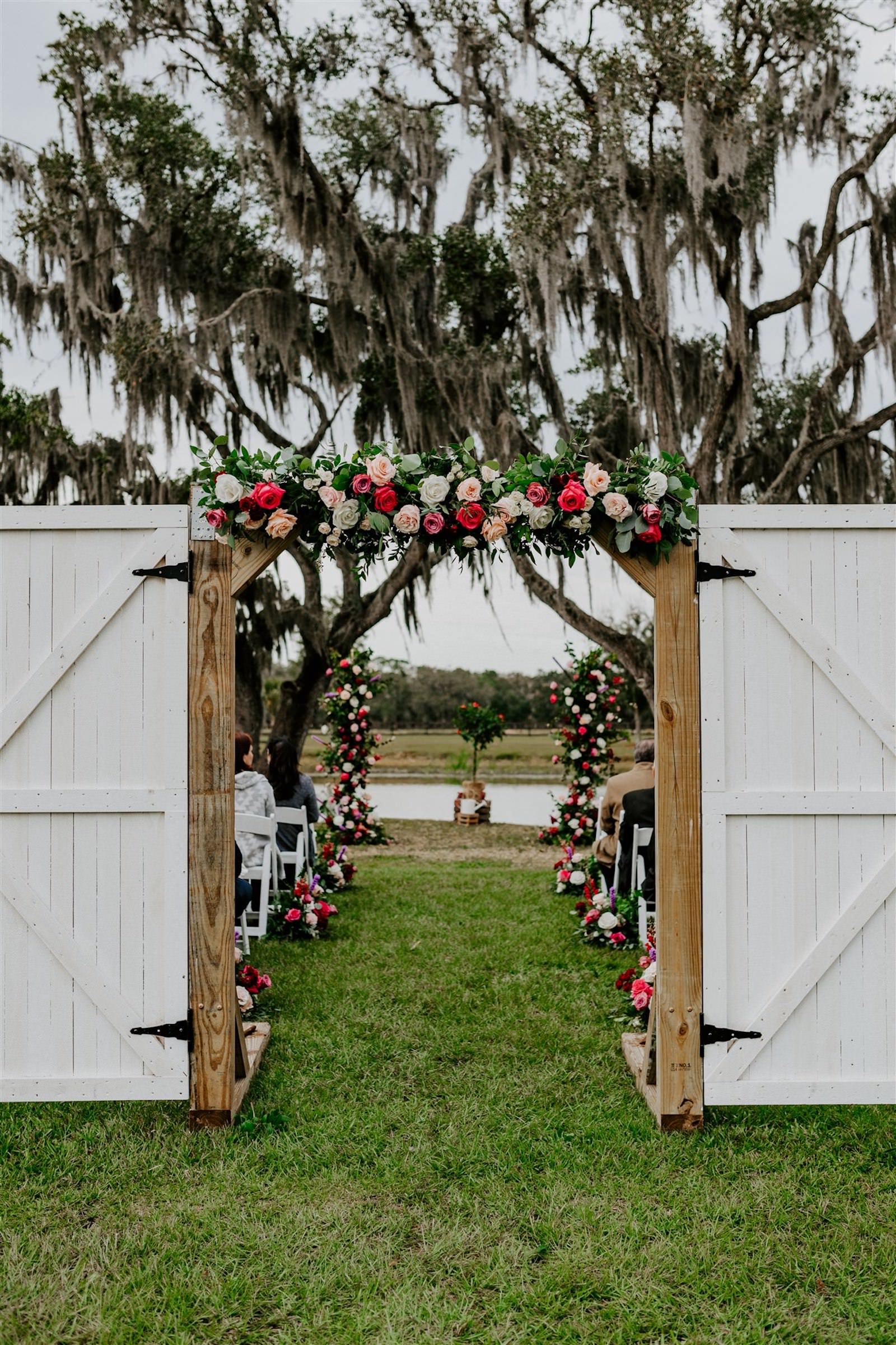 Rustic Tampa Outdoor Oak Tree Wedding Ceremony with White Garden Chairs | Wedding Barn Doors Entrance with Floral Garland | Tampa Wedding Florist Monarch Events and Designs | Deep Red Maroon Burgundy Roses and Blush Pink and White Roses with Eucalyptus Greenery