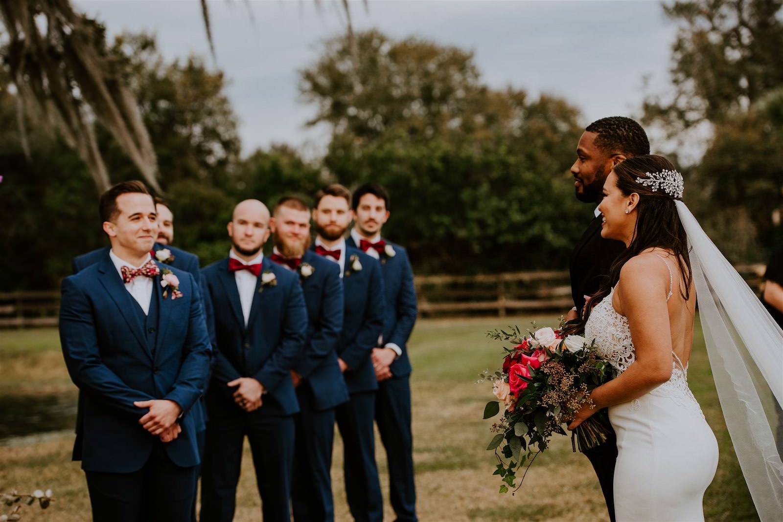 Bride Walking Down Aisle with Father during Outdoor Rustic Tampa Wedding | Groom and Groomsmen Wearing Navy Blue Suits with Red Bow Ties