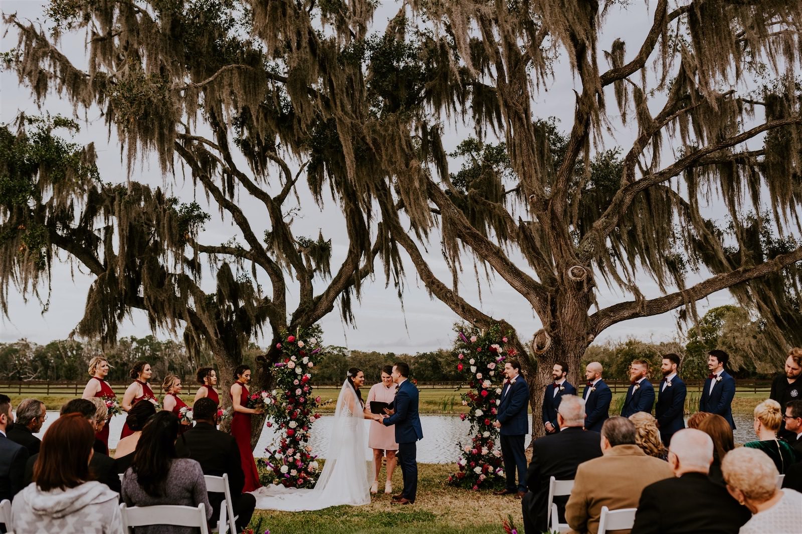 Rustic Tampa Outdoor Oak Tree Wedding Ceremony Backdrop Stands with Floral Garland | Tampa Wedding Florist Monarch Events and Designs | Deep Red Maroon Burgundy Roses and Blush Pink and White Roses with Eucalyptus Greenery | Groom and Groomsmen Wearing Navy Blue Suits | Maroon Burgundy Bridesmaids Dresses