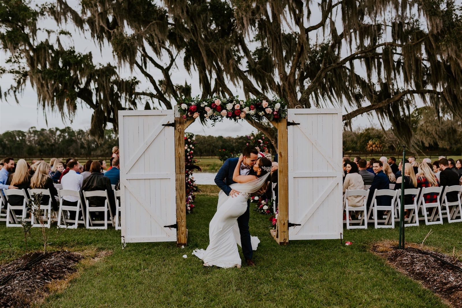 Bride and Groom Ceremony Exit Dip Kiss | Rustic Tampa Outdoor Oak Tree Wedding Ceremony with White Garden Chairs | Wedding Barn Doors Entrance with Floral Garland | Tampa Wedding Florist Monarch Events and Designs | Deep Red Maroon Burgundy Roses and Blush Pink and White Roses with Eucalyptus Greenery