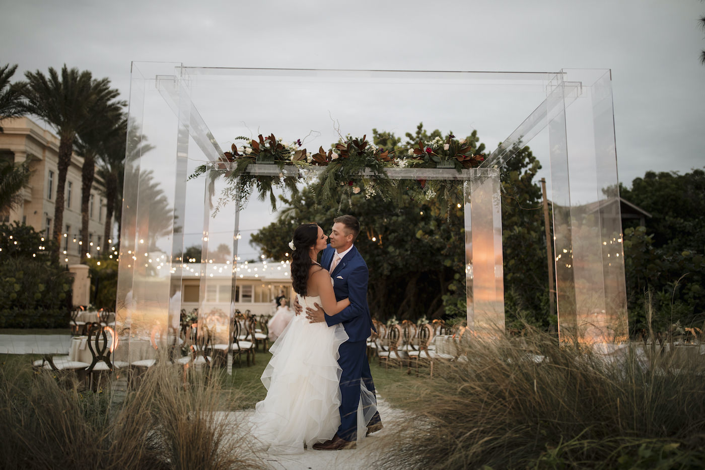 Bride and Groom Outdoor Portrait Sarasota Beach Wedding Ceremony on Siesta Key | Groom in Navy Blue Suit with Blush Pink Neck Tie | Horsehair Trim Tiered Organza Tulle Ballgown Bridal Gown with Sweetheart Neckline and Lace Bodice | Sarasota Wedding Dress Shop Truly Forever Bridal | Acrylic Four Post Arbor Backdrop with White and Deep Red and Blush Pink Floral Arrangement Garland with Eucalyptus Greenery