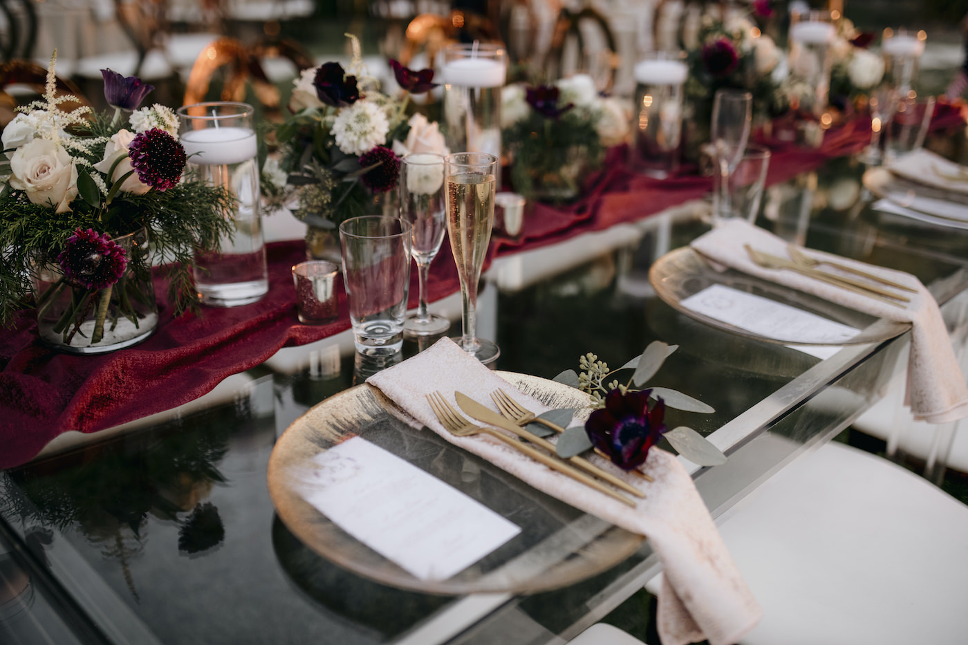 Outdoor Sarasota Beach Neutral Gold and Champagne Wedding Reception Table with Gold Glass Charger Plates and Gold Flatware and Red Velvet Table Runner | Gold Geometric Glass Frame Table Number | Gold Compote Vase Wedding Centerpiece with Floating Candles and White and Maroon Flowers and Greenery
