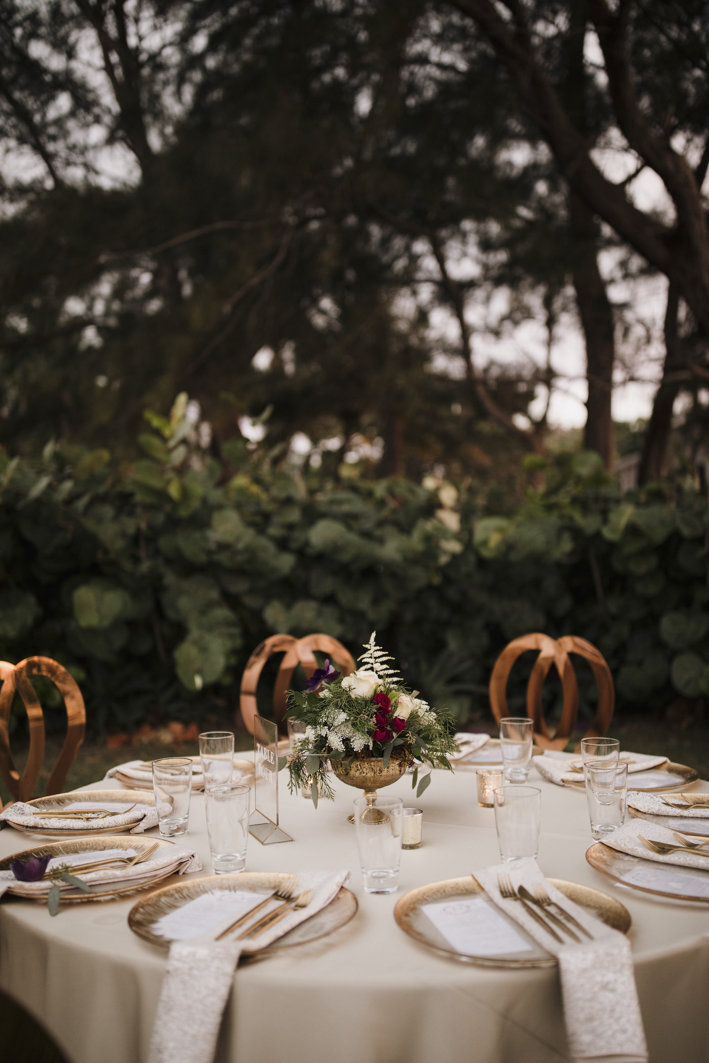 Outdoor Sarasota Beach Neutral Gold and Champagne Wedding Reception Table with Gold Glass Charger Plates and Gold Flatware and Gold King Louis Chairs | Gold Geometric Glass Frame Table Number | Gold Compote Vase Wedding Centerpiece with White and Red Flowers and Greenery