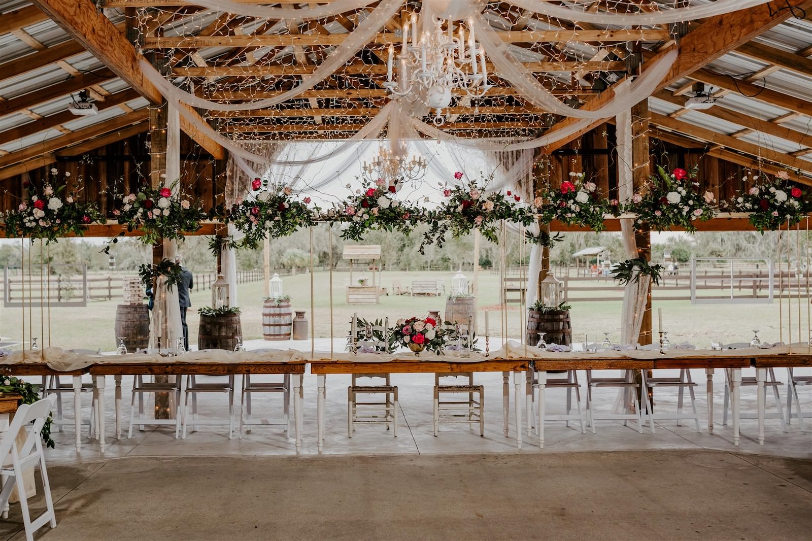 Rustic Barn Tampa Wedding with Suspended Ceiling Floral Arrangements with Blush Pink and Deep Red Burgundy Roses and Greenery | Wood Farm Tables with Champagne Linen Runners and Taper Candlestick Candles | Tampa Wedding Florist Monarch Events and Designs
