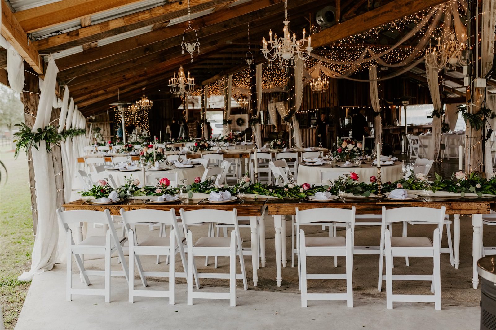 Rustic Barn Tampa Wedding with Twinkle Lights and Chandeliers | Garland Centerpieces with Blush Pink and Deep Red Burgundy Roses and Greenery | Wood Farm Tables with Champagne Linen Runners and Taper Candlestick Candles and White Garden Chairs | Tampa Wedding Florist Monarch Events and Designs
