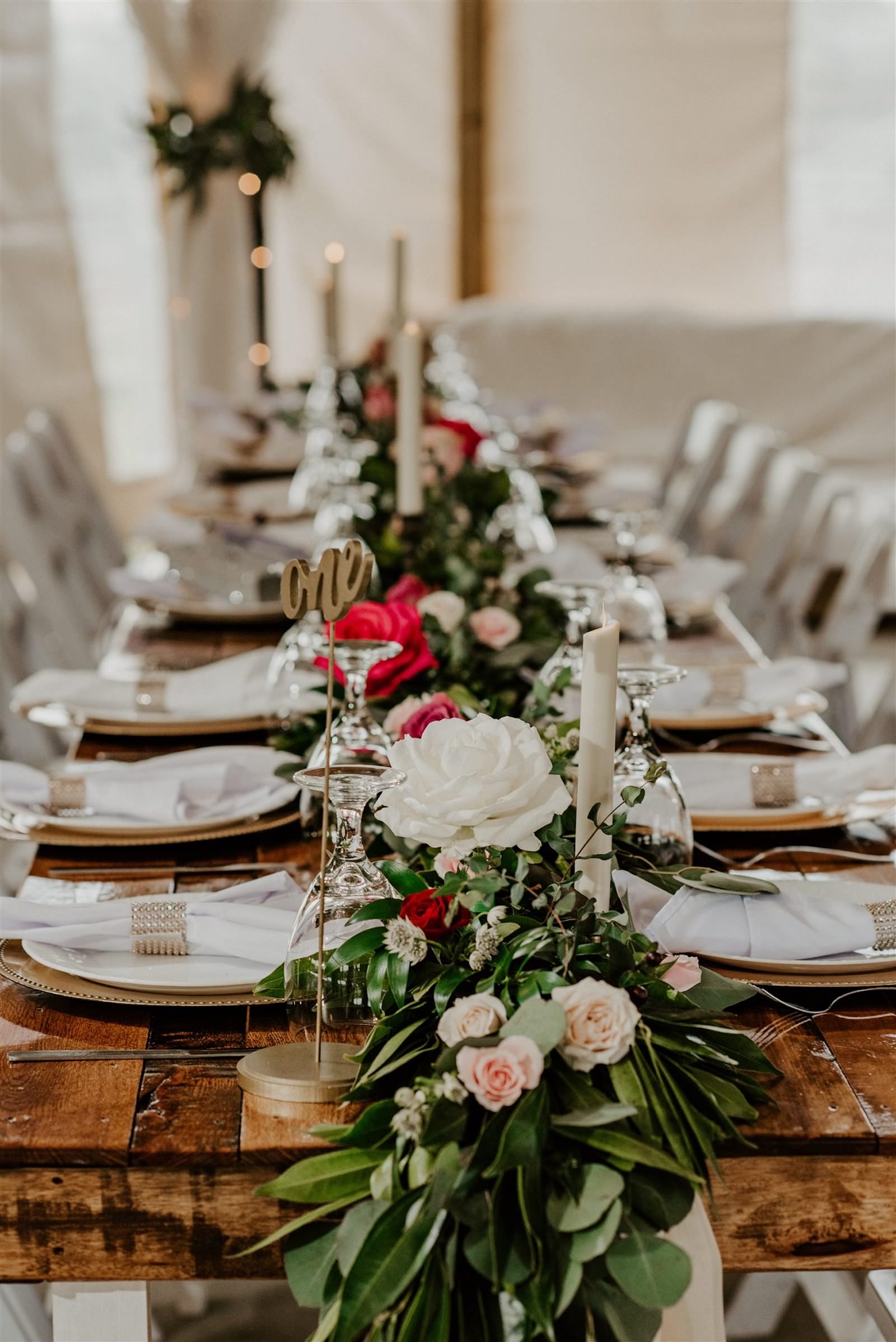 Rustic Barn Tampa Wedding Garland Centerpieces with Blush Pink and Deep Red Burgundy Roses and Greenery | Wood Farm Tables with Champagne Linen Runners and Taper Candlestick Candles and White Garden Chairs | Tampa Wedding Florist Monarch Events and Designs