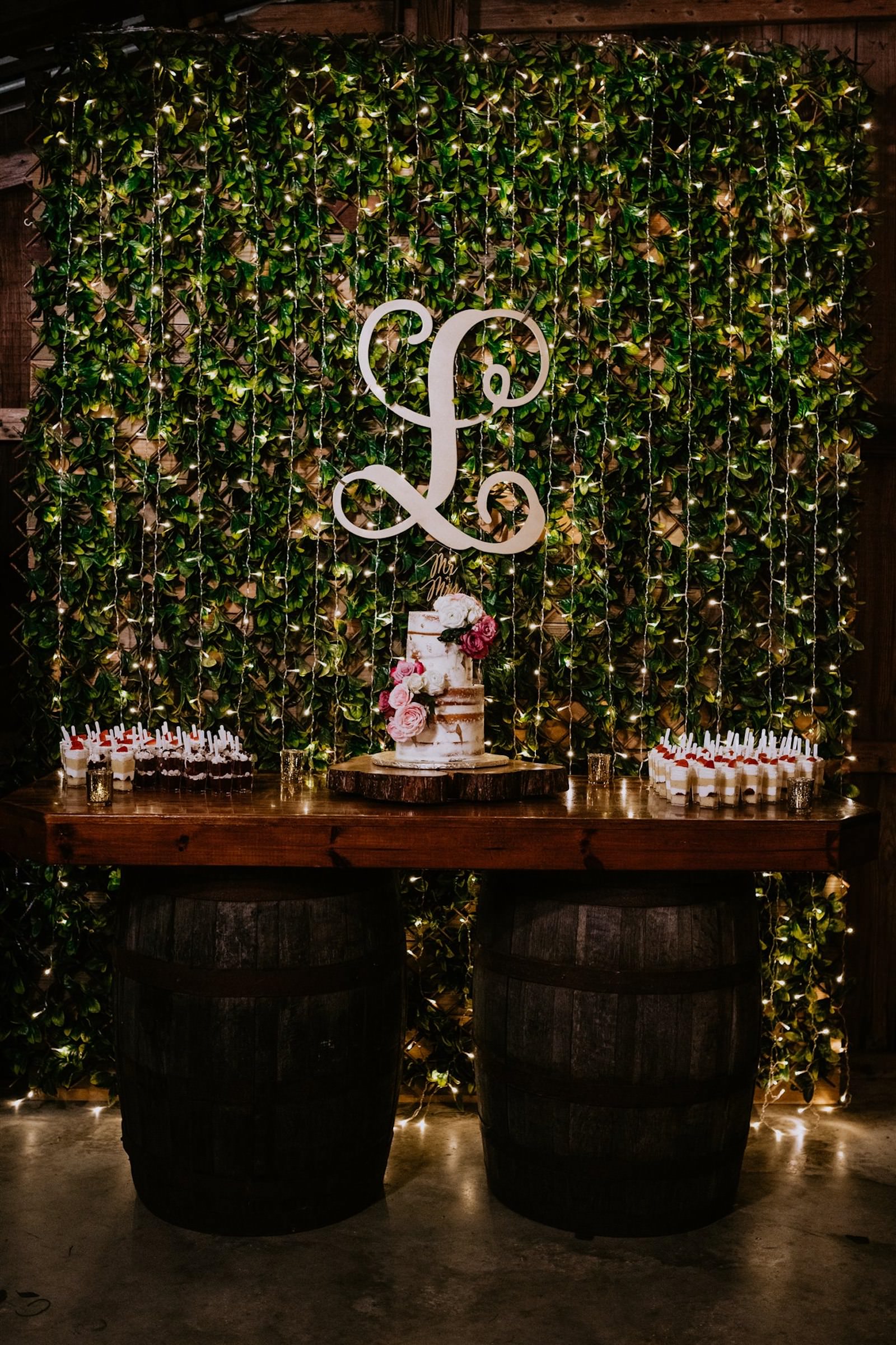 Wood Barrel Wedding Cake Table with Greenery Wall Backdrop and Twinkle Lights and Monogram Sign | Two Tier Naked Cake with Fresh Garden Rose Peony Flower Accents | Mini Dessert Shot Parfaits Wedding Dessert Table